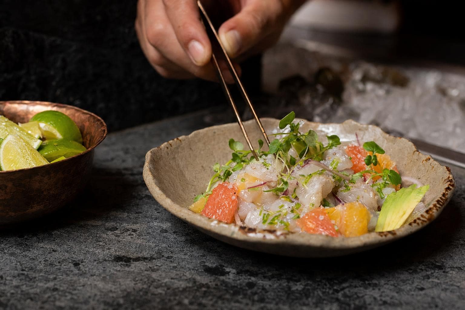 Chef garnishing ceviche with bronze tweezers at the raw bar — citrus, micro herbs, sea salt on a handmade ceramic bowl